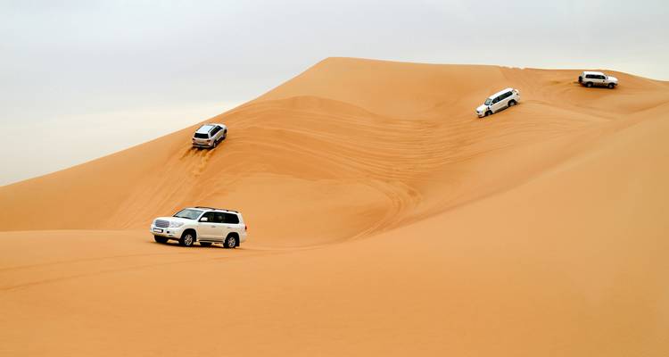 Quatre véhicules 4x4 tracent des chemins le long d'une vaste dune dorée lors d'un safari désertique.