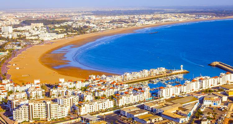 Hoge luchtopname van Agadir's halvemaanvormige strand, jachthaven en uitgestrekte stad omgeven door blauwe oceaan.