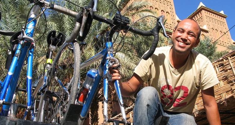 Un cycliste souriante s'accroupit à côté de vélos de randonnée bleus devant une casbah traditionnelle en adobe marocaine et des palmiers.