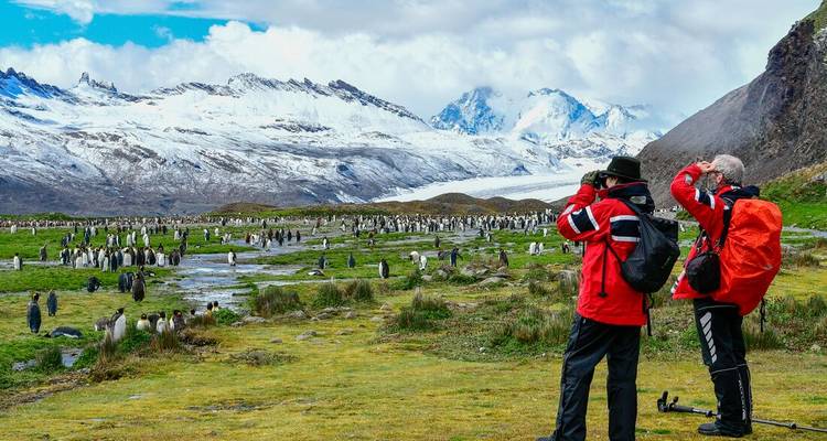 Two travelers with cameras scan a broad field filled with penguins against a backdrop of glaciers and peaks