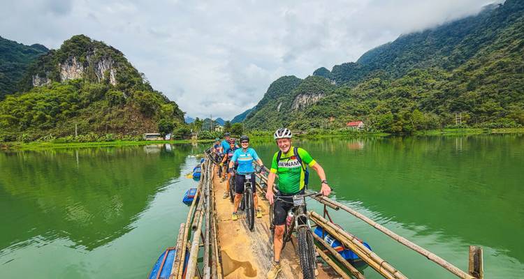 Cyclists push bikes across a narrow bamboo bridge over emerald water, flanked by karst mountains.
