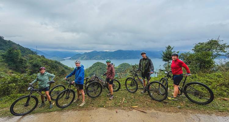 Five cyclists line up on a mountain road with a sweeping lake and forested hills below.