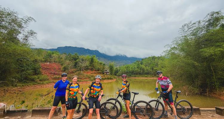 Cyclists stop by a small pond with mist-covered mountains and bamboo forest around.