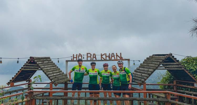 Cyclists in matching green jerseys stand below a wooden sign reading Ho Ba Khan overlooking a lake.
