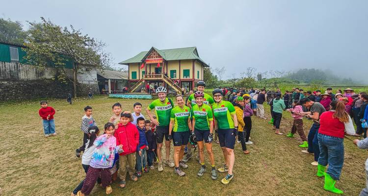 Cyclists mingle with a large group of village children in front of a traditional stilt house on a foggy morning.