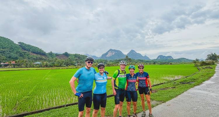 Cyclists pose beside vibrant green rice paddies with limestone peaks in the distance after rainfall.
