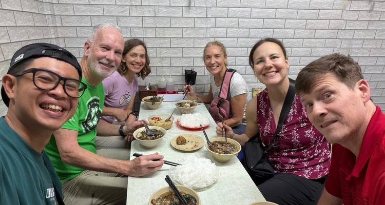 Group of travelers enjoy bowls of Vietnamese noodles together inside a small tiled eatery.