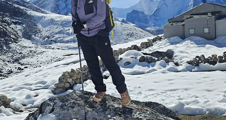 Een trekker staat op een besneeuwde rotsblok met wandelstokken, ruige berglandschap op de achtergrond.