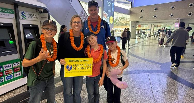 Family wearing marigold garlands smiling with a welcome sign inside Kathmandu airport arrival hall.