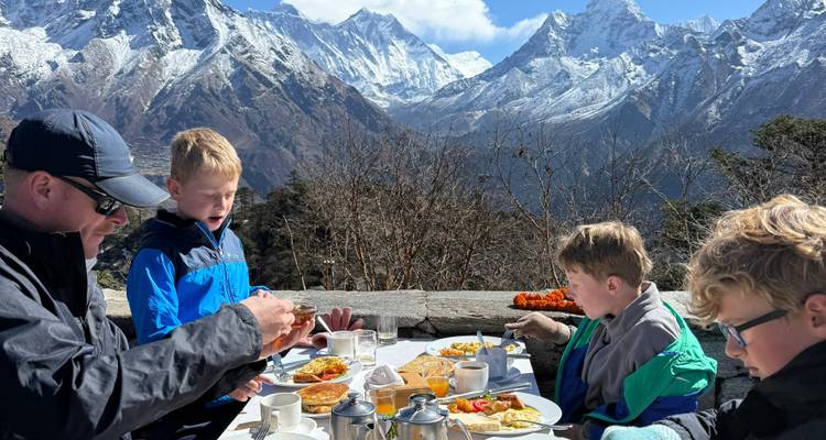 Family enjoying outdoor breakfast with snow-covered Himalayan peaks towering behind.
