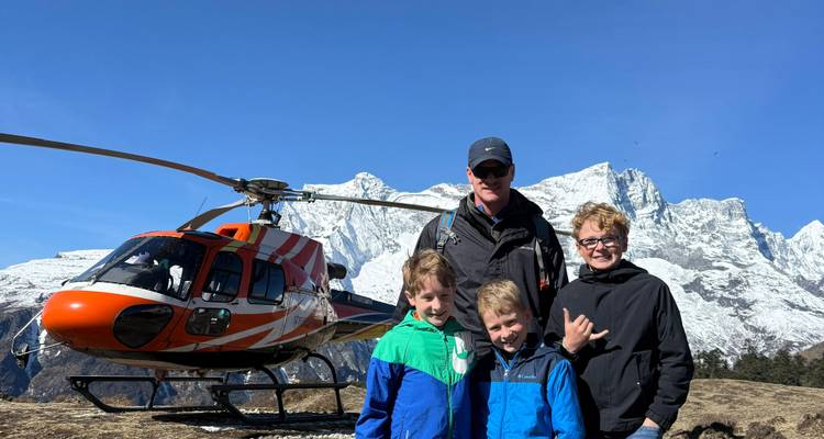 Family poses beside bright orange helicopter with snowy Himalayan peaks in the background.
