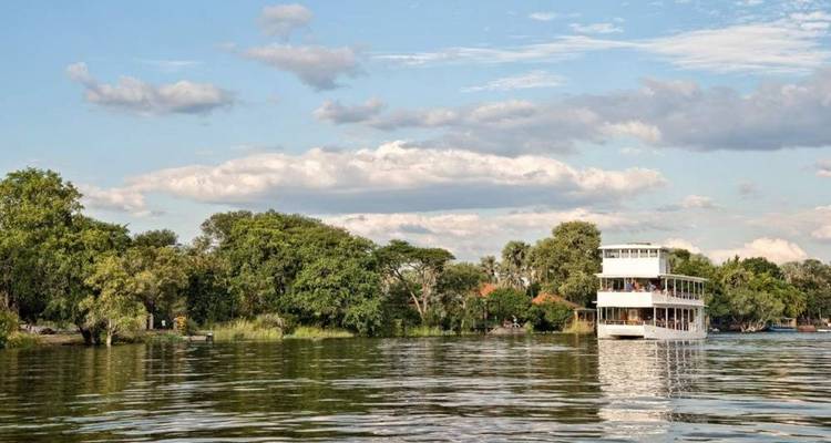 Weißes zweistöckiges Flusskreuzfahrtschiff gleitet entlang eines breiten Flusses, gesäumt von grünen Bäumen unter einem blauen Himmel mit Wolken.