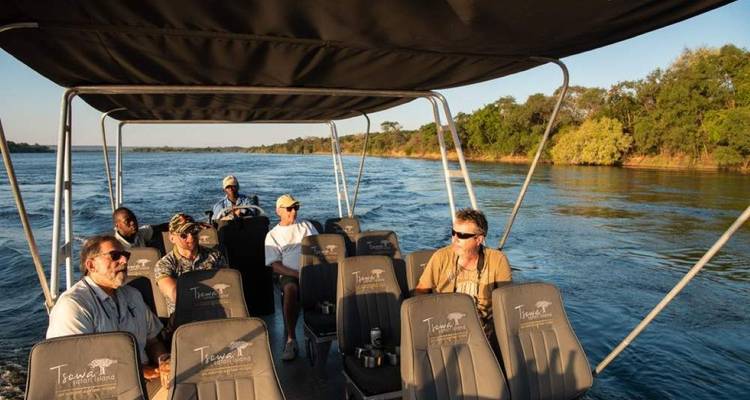 Kleines Safari-Boot mit Touristen fährt durch schnell fließenden Fluss, gesäumt von Grün in warmem Licht.