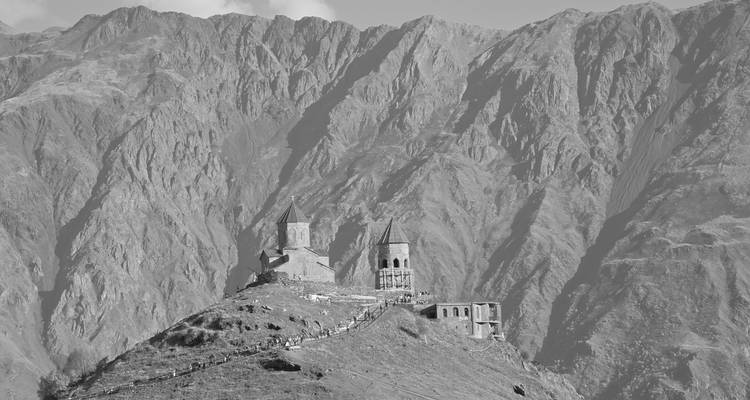 Zwart-wit close-up van de Gergeti Drievuldigheidskerkcerk tegen dramatische rotswanden.