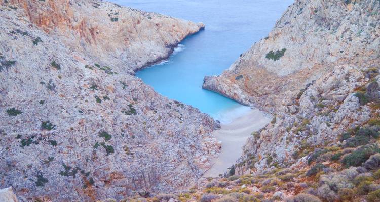 Smalle rotsachtige kloof die uitkomt op een verborgen baaitje met helder blauw water en een klein zandstrand.