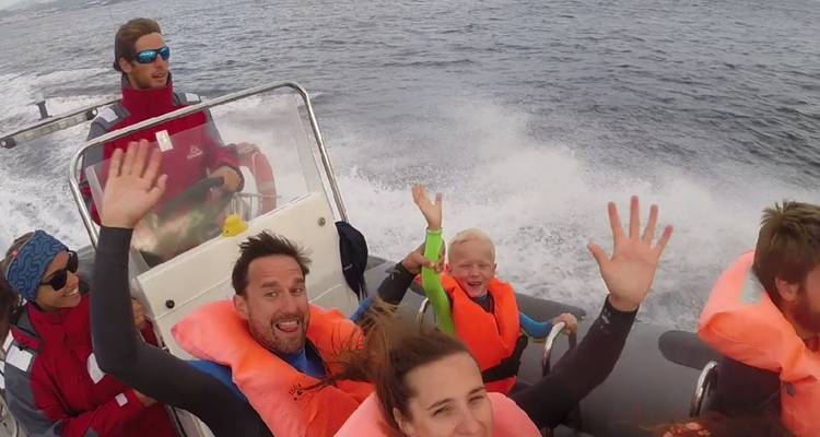Smiling passengers in life jackets waving on a fast inflatable boat with sea spray behind.