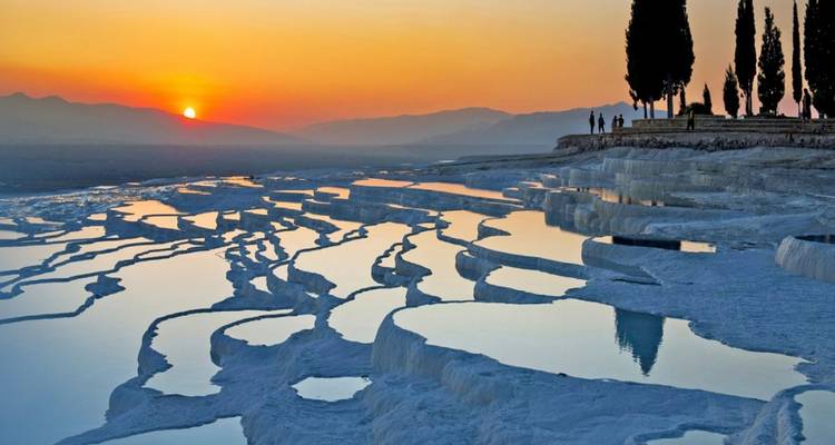 Pamukkale-Travertinterrassen, die die lebendigen Farben des Sonnenuntergangs widerspiegeln, mit silhouettierten Zypressen am Horizont