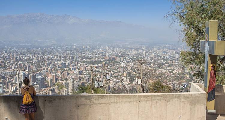 Viajera con mochila contempla una vasta vista de la ciudad desde una terraza en la cima de una colina con un monumento de cruz.