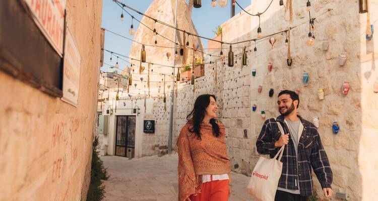 Lächelndes Paar spaziert durch eine schmale Steingasse, die mit hängenden Lichtern und Flaschen in einer kappadokischen Stadt geschmückt ist.