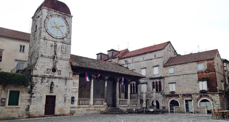 Plaza del pueblo en Trogir con torre del reloj de piedra, banderas croatas y mesas de café vacías.