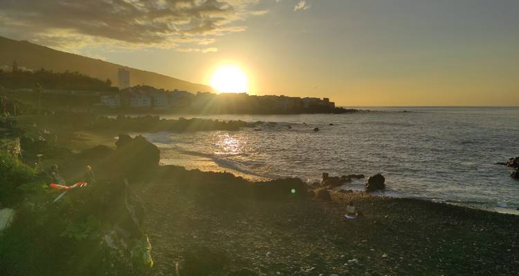 Sun sets over a rocky shoreline casting golden light on gentle waves and silhouetted village