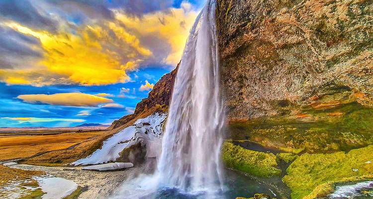 Tall waterfall cascades over a cliff as dramatic golden clouds color the Icelandic sky at dusk.
