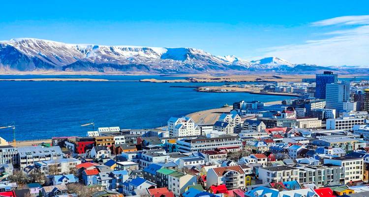Panoramic aerial of colorful Reykjavik houses along the waterfront with snow-topped mountains in the background.