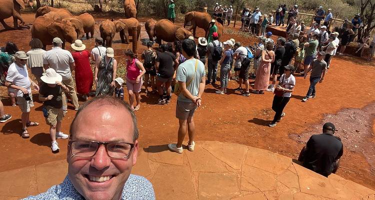 Tourist takes a selfie above a red-soil enclosure where numerous visitors observe baby elephants