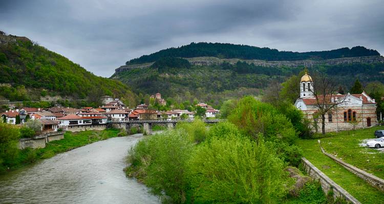 Rivierdorp, stenen brug en beboste heuvels onder grijze lentelijke luchten in Bulgarije.
