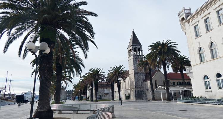 Palmengesäumte Promenade mit historischem Glockenturm und Wasserfront-Gebäuden im sanften Licht.