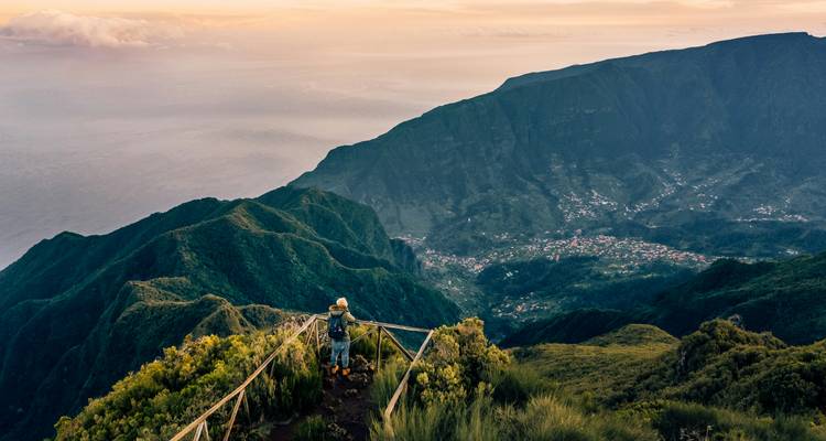 Een wandelaar staat op een omheind uitkijkpunt terwijl gouden licht over weelderige groene bergen spoelt die afdalen naar een kustdal ver beneden.