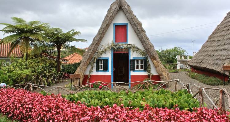 Een traditioneel A-frame Santana huisje met helderrode basis en blauw omlijste ramen staat te midden van levendige bloembedden en weelderig gebladerte.