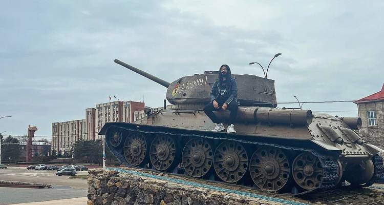 Jonge man zittend bovenop een historische Sovjet-tank tentoongesteld op een stadsplein op een bewolkte dag.