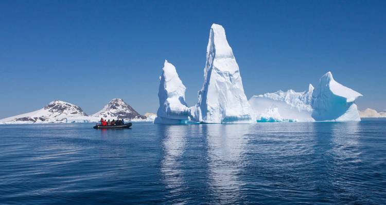 A small Zodiac boat of tourists floats beside towering sculptural icebergs under a clear blue Antarctic sky.