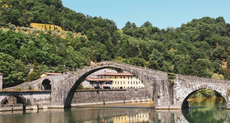 Un élégant pont en arc de pierre de style médiéval enjambe une rivière calme avec des collines boisées et des maisons de village à proximité.
