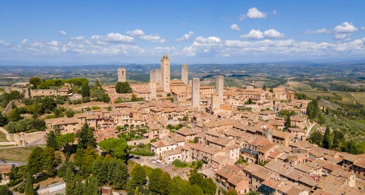 Vue aérienne d'une ville médiévale perchée sur une colline, couronnée par plusieurs tours en pierre au milieu de la campagne toscane vallonnée.