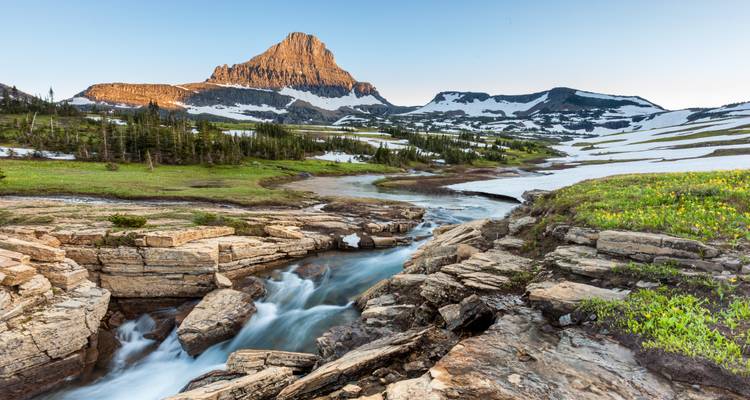 Crystal stream rushing through rocky alpine meadow beneath towering peak at sunrise