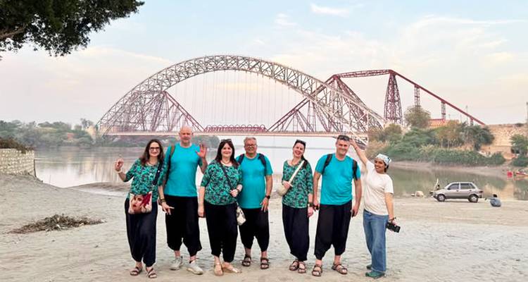 Groupe portant des chemises turquoise assorties pose sur une berge sablonneuse avec les ponts en fer de Sukkur en arrière-plan.