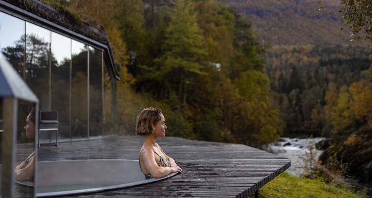 Mujer relajándose en piscina exterior humeante empotrada en terraza de madera junto a cabaña de cristal y río