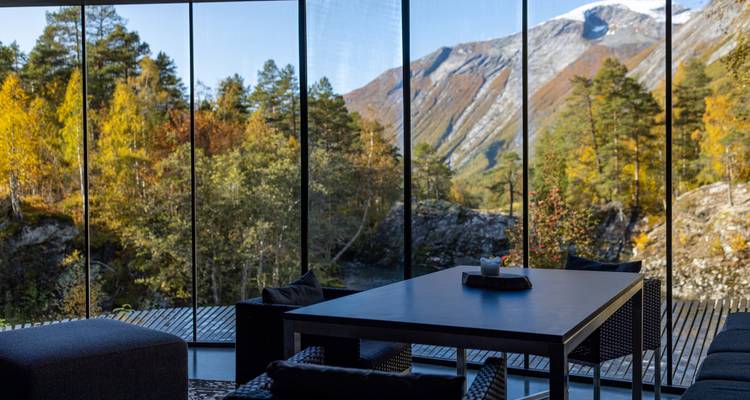 Área de estar con paredes de cristal panorámicas con vista al bosque otoñal y montañas cubiertas de nieve