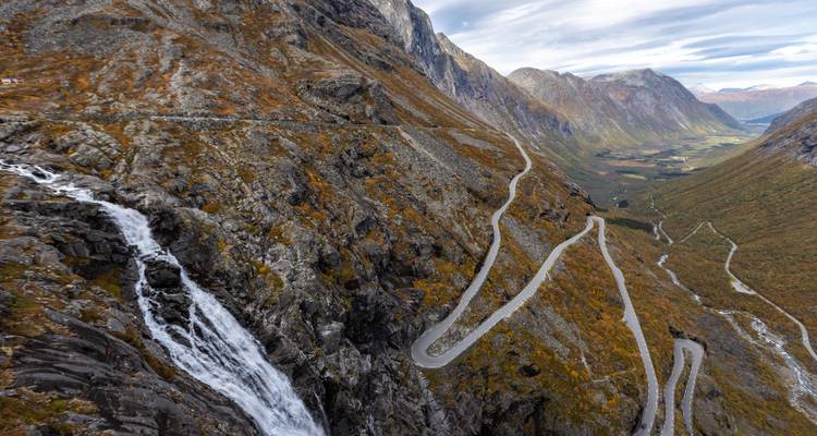 Toma aérea dramática de carretera serpenteante de montaña con cascada precipitándose por un acantilado hacia el valle