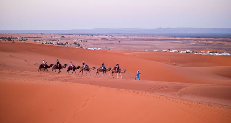 Een Berberse gids leidt een karavaan kamelen met reizigers over oranje duinen bij zonsondergang.