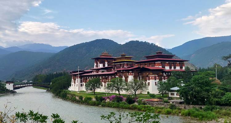 Large riverside dzong with red and white walls set against green hills and flowing water.