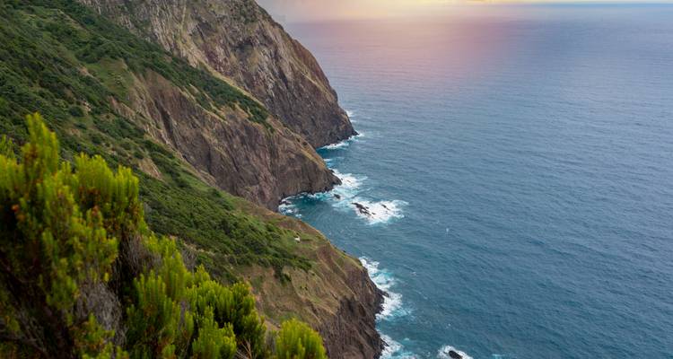 Acantilado verde empinado se precipita hacia el Atlántico con olas espumosas contra las rocas al atardecer en la costa de Madeira.