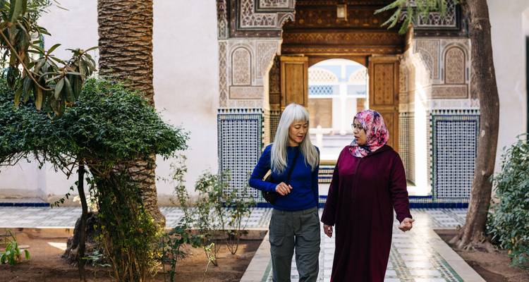 Two women converse while strolling through a richly tiled courtyard of a Moroccan palace.