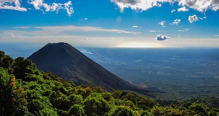 A conical volcanic peak rises above lush jungle with distant ocean views under a bright sky.