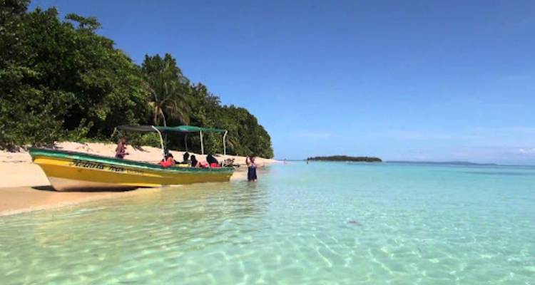 A small boat with tourists anchors in crystal-clear shallow water beside a pristine sandbar.