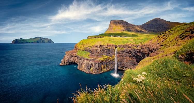 De iconische Múlafossur waterval stort zich van groene kliffen in de Noord-Atlantische Oceaan naast een klein dorp op de kliftop en zeerotsen.