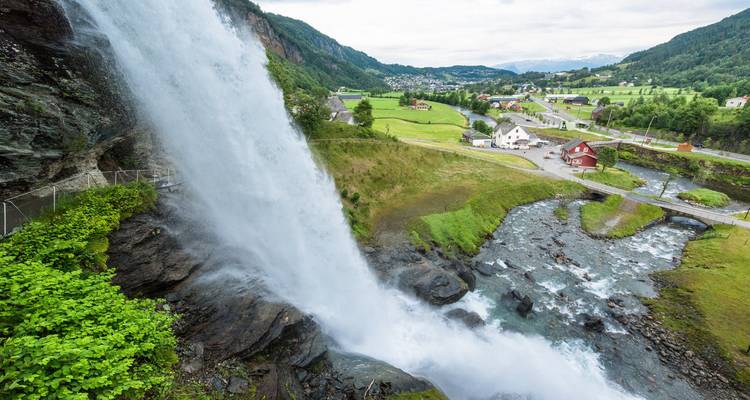 Krachtige waterval die naar beneden stort naast een landelijk dorp met velden, huizen en een kronkelende beek eronder.
