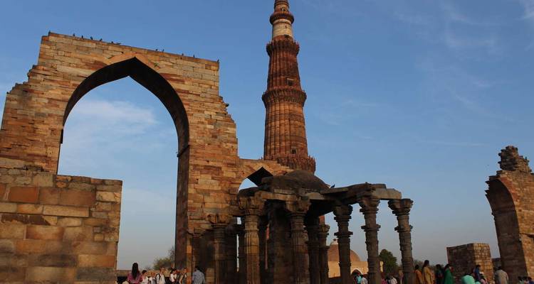 Ancient stone arch and Qutub Minar tower rising above temple ruins under blue sky.